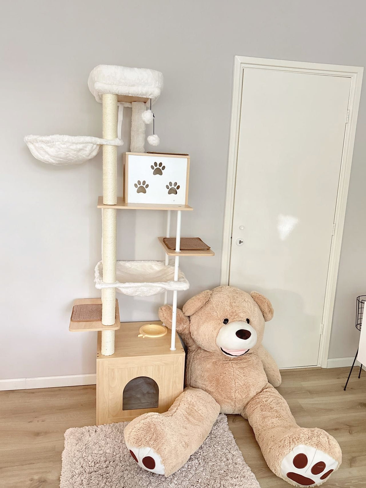 Cat tree with wooden steps and platforms, next to a large teddy bear on a wooden floor.